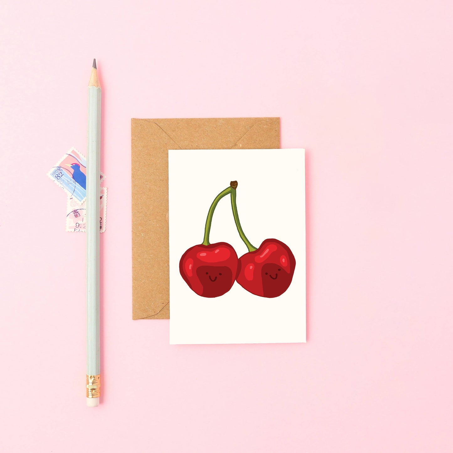 A greeting card with a hand-drawn illustration of two smiling cherries, titled 'Mini Cherries Card'. The card is A7 sized, comes with a brown Kraft envelope, and is blank inside.