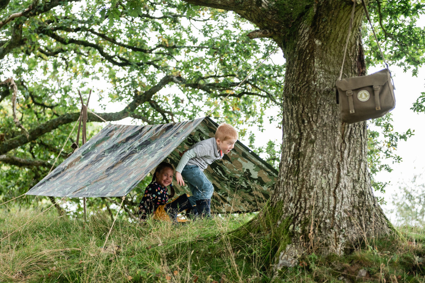 Two children enjoying themselves in their den. The den kit bag hangs on a nearby tree.