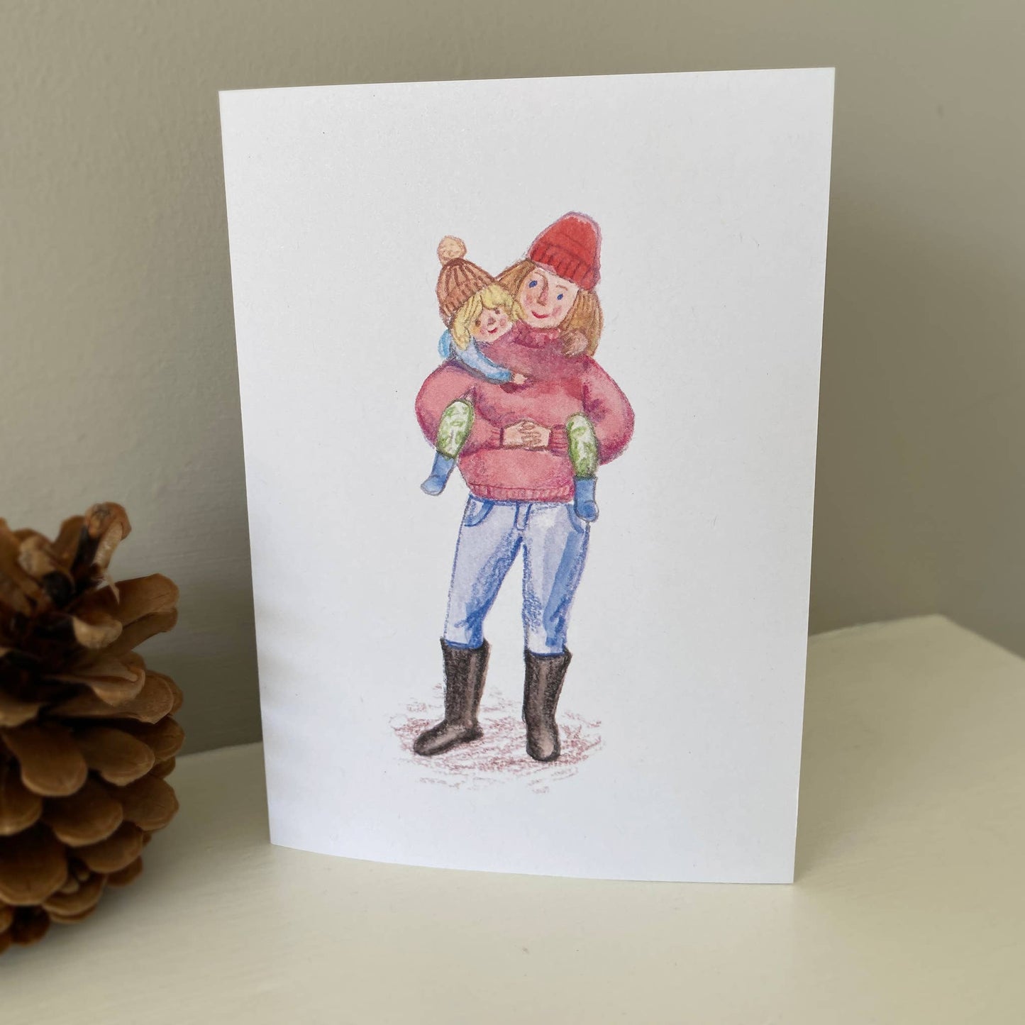 Inspired by muddy walks in the forest with her children, this card is illustrated by Juliet Thomas Doodles. This card shows a Mum, giving her child a piggyback out for a walk in their winter woollies.