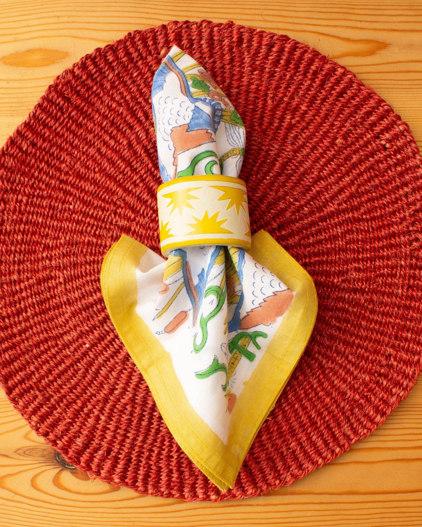 Hand wood block printed organic cotton napkins made in Jaipur, India. Add a touch of joy to your dining table with our beautiful set of four organic cotton napkins. Featuring AARVEN's signature 'Kingfisher' print. Block printed with different colourful borders (green, blue, coral and yellow) onto a white organic cotton background. Shown with a yellow and white napkin ring against an orange circular placemat.