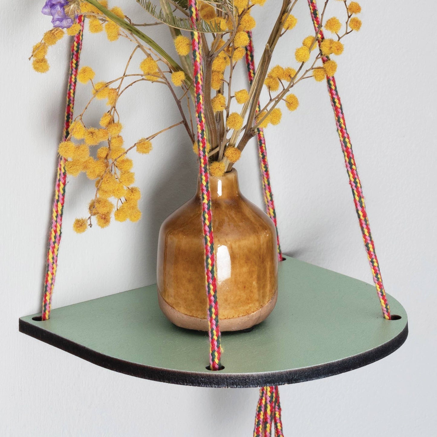 Close up of a green wash ‘plant shelf’ that rests neatly against the wall. Colourful twine contrasts nicely with the shelf to make a standout piece. Brass plated hoops and crimp bars finish the look No assembly required. Shown against a white wall, with a small vase of dried flowers.