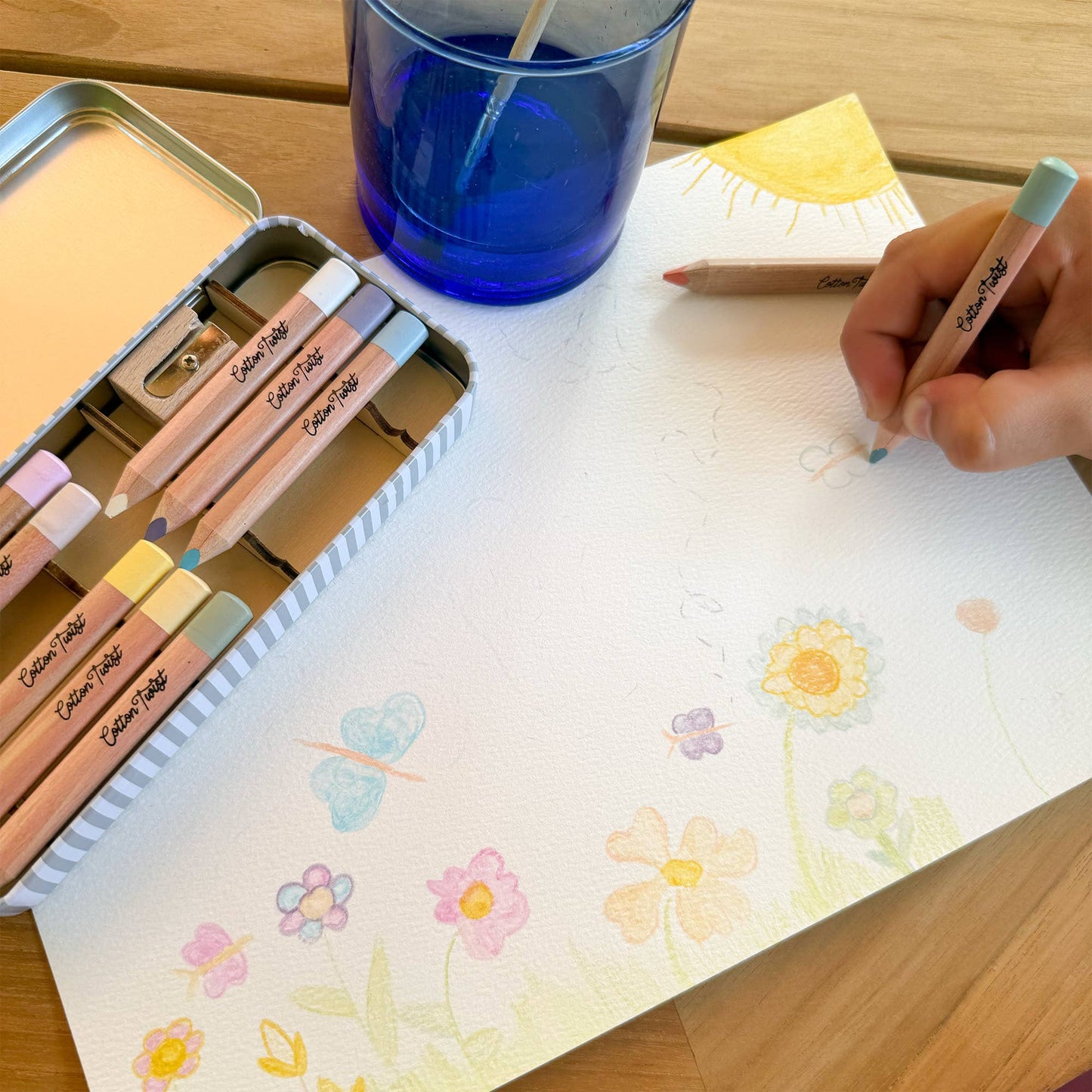 Colouring with pastel-coloured pencils on a piece of paper with floral designs, next to a blue glass and pencil sharpener.