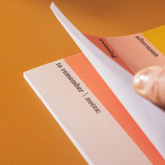 A Daily Planner Pad with pastel colour blocks in pink, orange, and yellow, with space for writing 'first thing', 'morning', 'afternoon', 'evening', and 'to remember' tasks, placed on a desk with a pen and decorative items in the background.