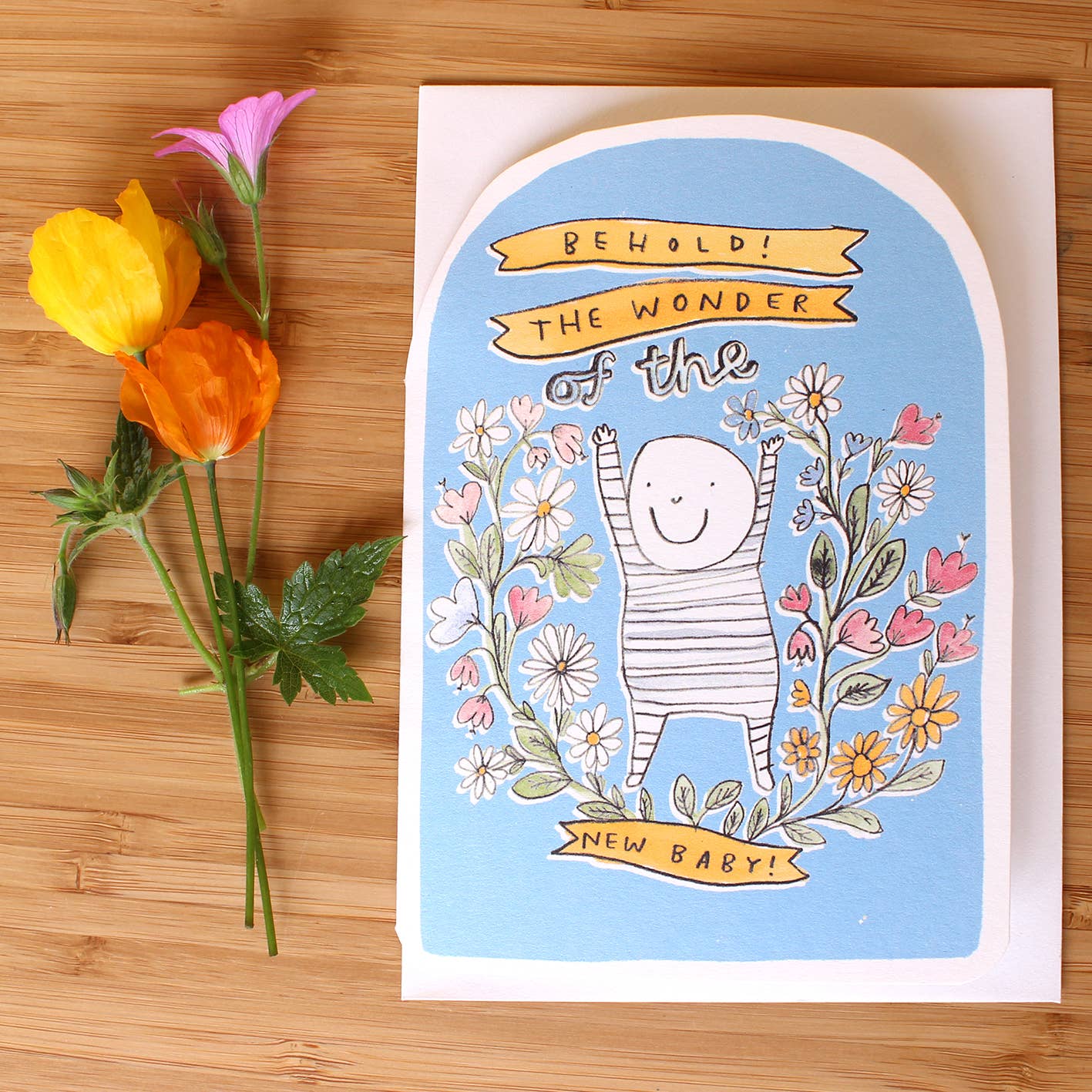 Close-up of a new baby greeting card titled 'Behold! The Wonder'. Features a black and white illustration of a smiling baby in a striped onesie, surrounded by pink and yellow flowers on a blue background. The card has a unique oval-shaped top.