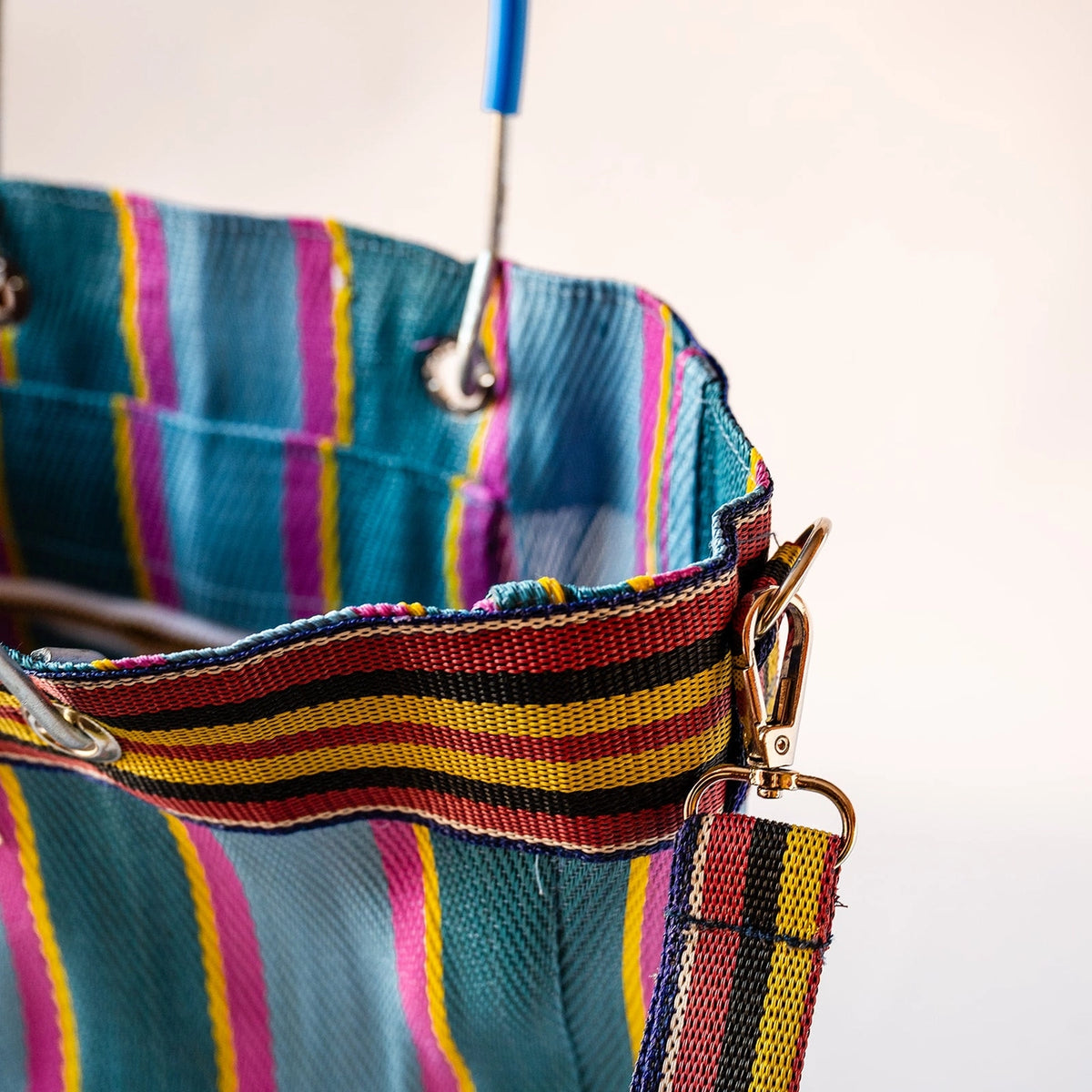 Close-up of a colorful striped fabric bag or tote with a woven strap, metal grommets, and gold-tone hardware against a plain light background.