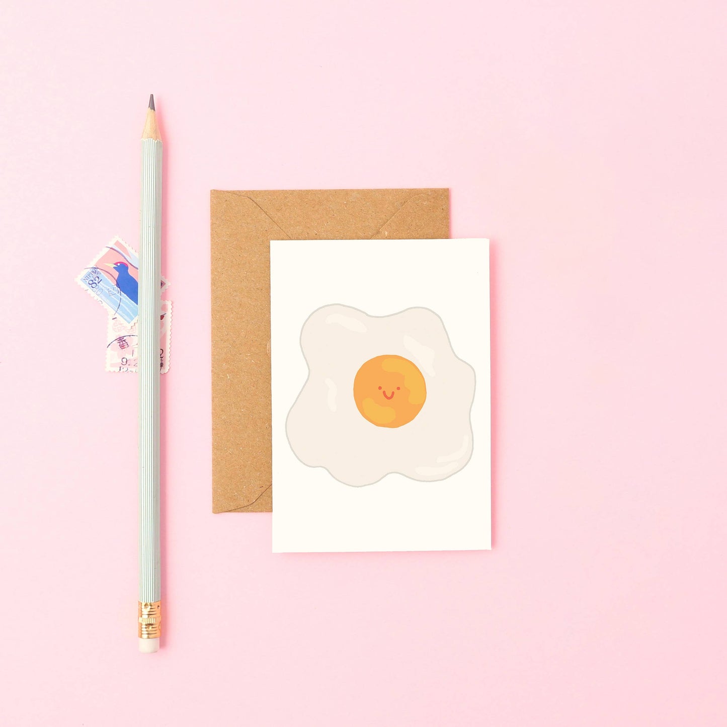 Card with a smiley face egg design on a pink background with a pencil and envelope.