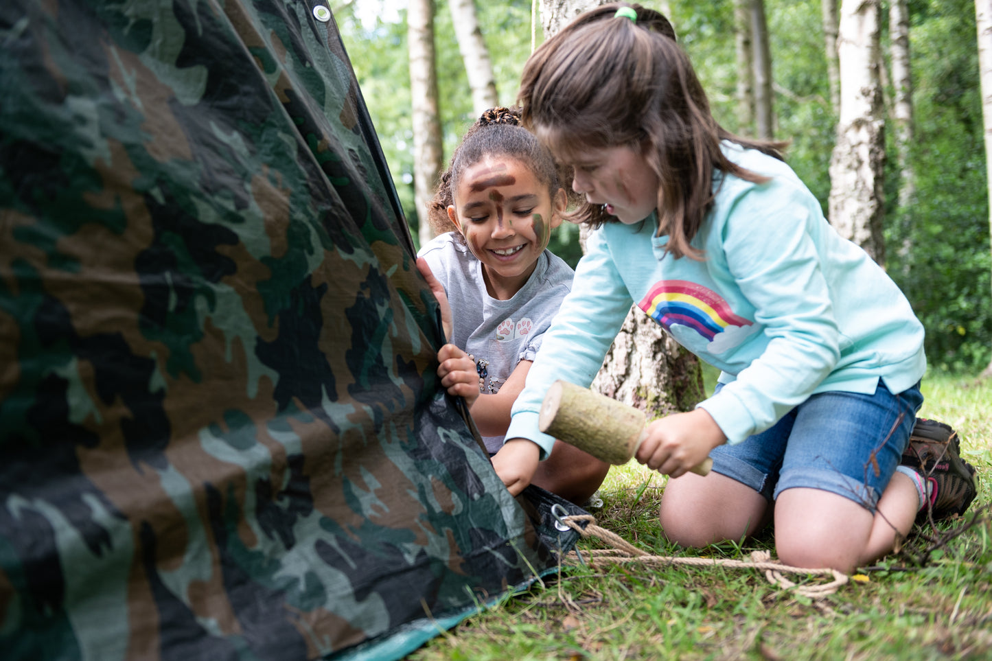 Two children help each other, bang the tent pegs into the ground with the wooden mallet. Both children have painted their faces with the camouflage face paint.