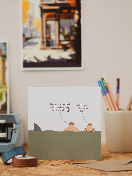 Close-up of Wild Swimming greeting card by Not at all Jack, showing cartoon swimmers and shark fin detail, sustainable.