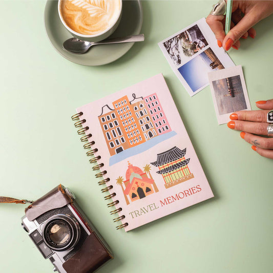 A travel memories journal with a hardcover featuring colorful illustrations of iconic landmarks, a camera, and a cup of coffee on a table. A person is holding a photo in the background.
