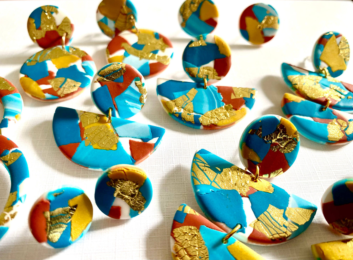 Large polymer clay statement earrings with turquoise, teal, white and terracotta colours, and splashes of gold leaf. Nickel Free. The earrings are in three parts a round section with a small odl hoop connecting it to a large semi circle and then another small gold hoop connecting it to a smaller semi circle. So the earring dangles below the ear lobe. Shown with other styles in the same pattern.