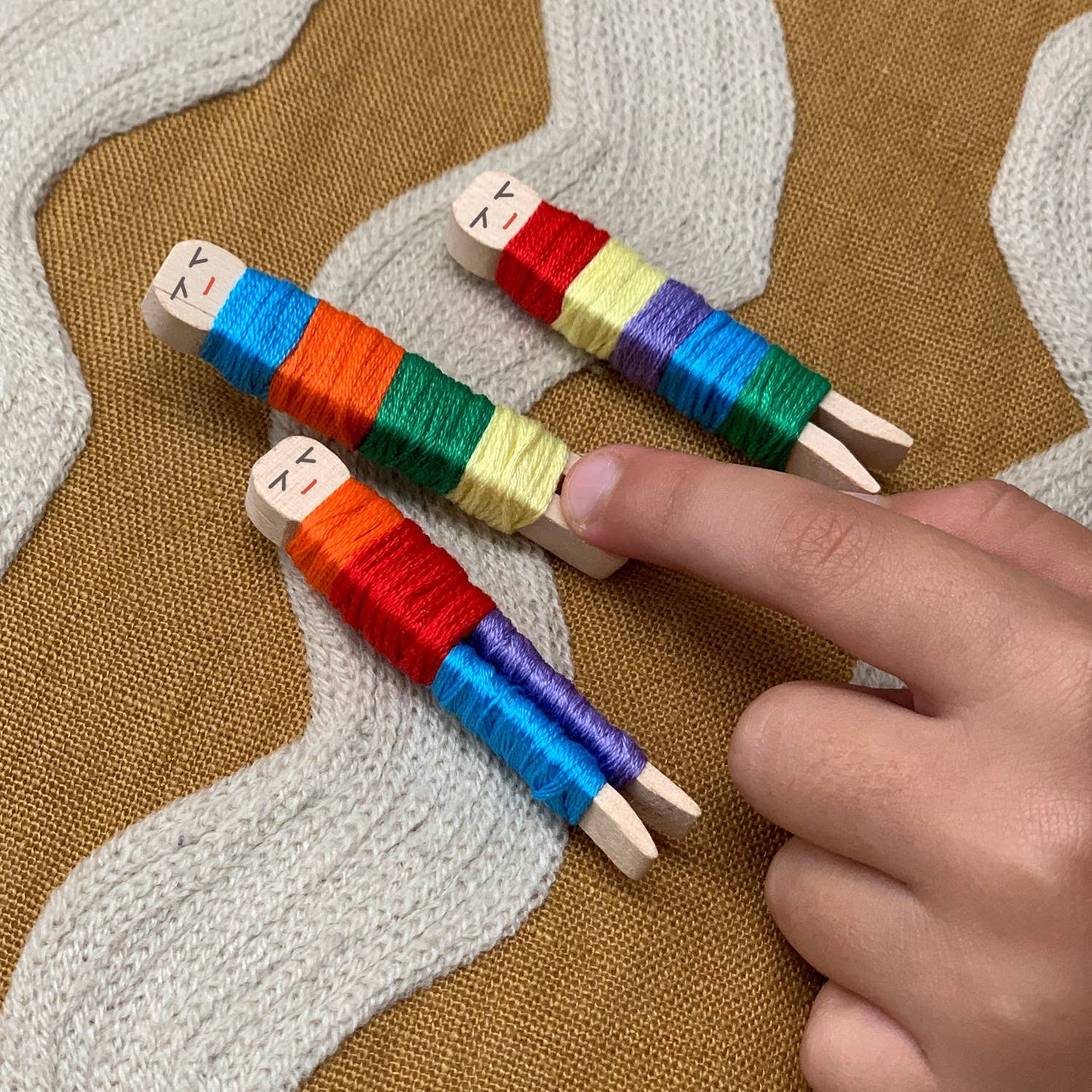 Colorful thread spools on wooden pegs with a hand interacting with them against a textured fabric background.