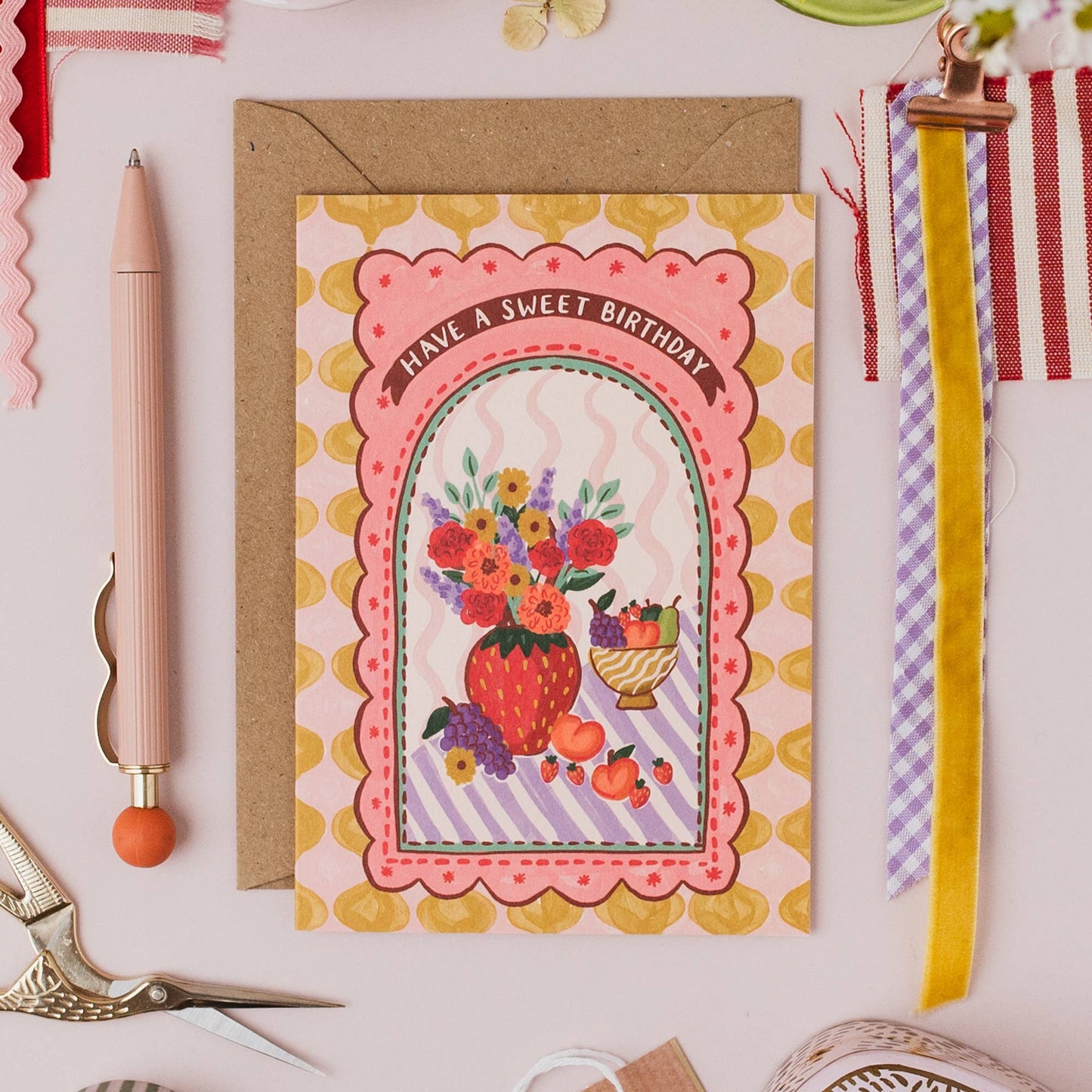 Hand drawn lettering reads 'Have a sweet birthday'. Part of the on trend Harlequin range, which features contemporary vases of flowers and fruit in a patterned tablescape. A6 size illustrated greeting card with tile pattern and vase of flowers and fruit illustration, printed onto premium textured card with a kraft envelope.