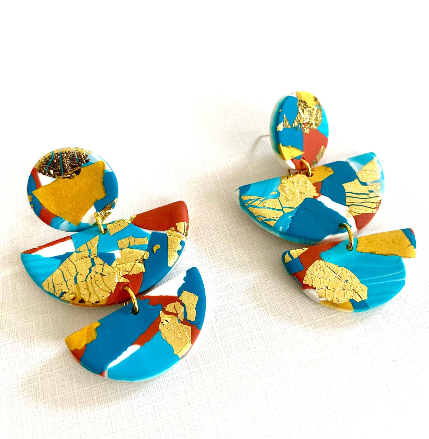 Large polymer clay statement earrings with turquoise, teal, white and terracotta colours, and splashes of gold leaf. Nickel Free. The earrings are in three parts a round section with a small odl hoop connecting it to a large semi circle and then another small gold hoop connecting it to a smaller semi circle. So the earring dangles below the ear lobe.