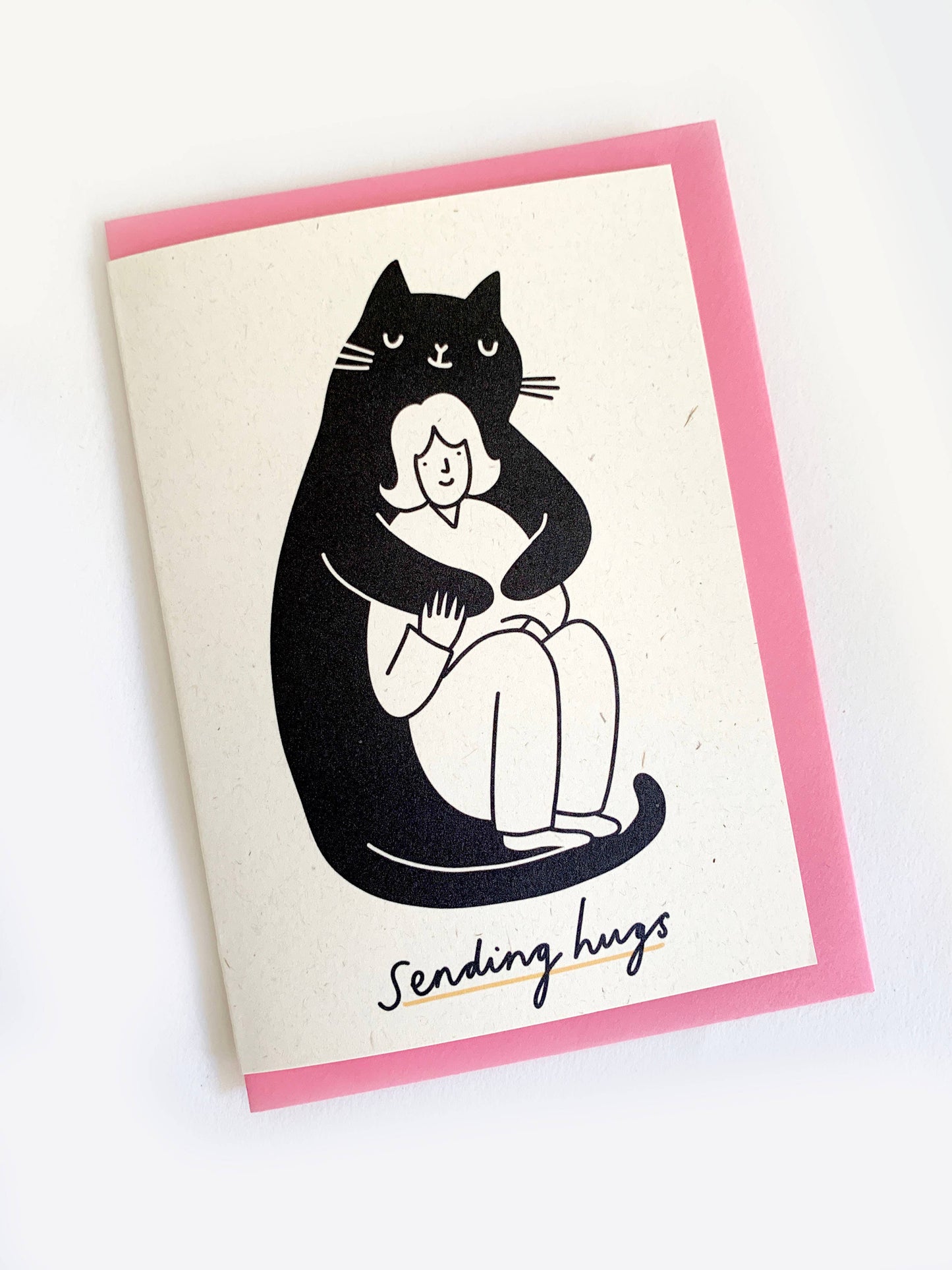 Black and white illustration of a huge black smiling cat cuddling a smaller smiling woman. with the words sending hugs at the bottom of the card.