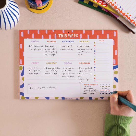A colourful A4 weekly planner pad with a weekly view layout from Monday to Sunday, with sections for important tasks, to-do lists, and a habit tracker. The planner is placed on a desk with stationery items around it.