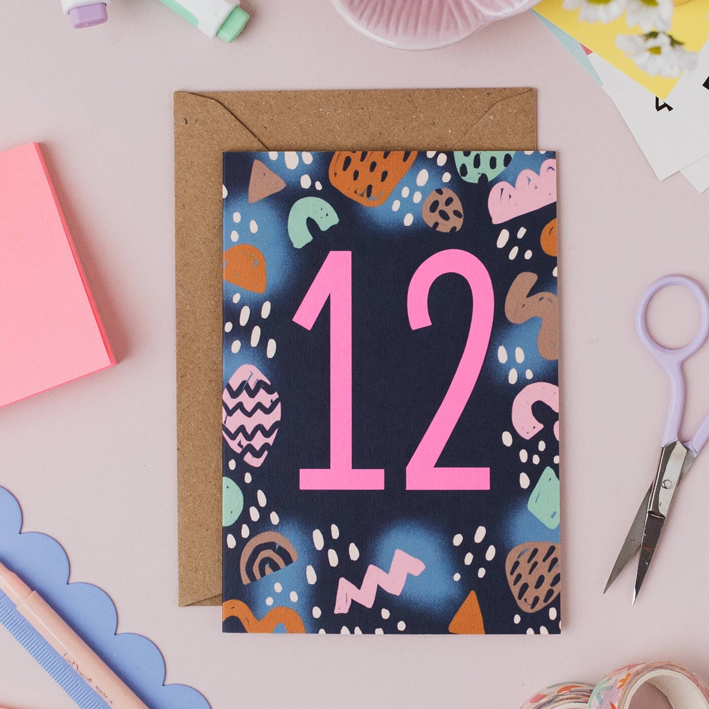 This on trend colour palette and neon pink number makes this milestone birthday card stand out on the shelf. Printed onto premium matt card with a kraft envelope. Illustrated greeting card with abstract shapes pattern and neon pink ink number 12.