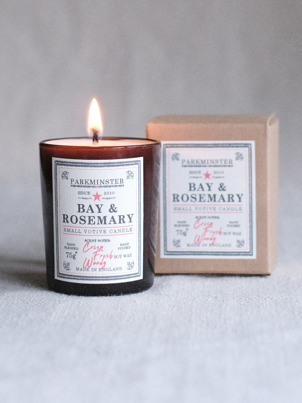 A lit amber glass candle jar labeled 'Bay & Rosemary' stands in front of its kraft paper box, also labeled 'PARKMINSTER' and 'Bay & Rosemary,' on a light gray textured surface.