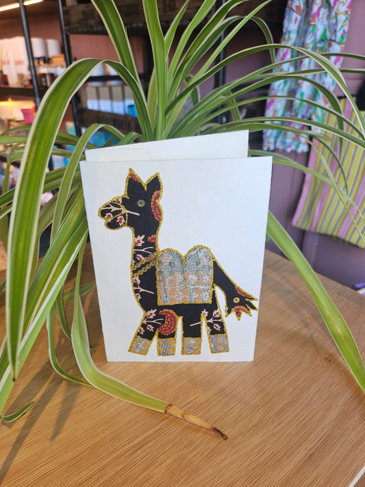 Decorative card with a camel design on a white background, placed on a wooden surface with a plant in the foreground.