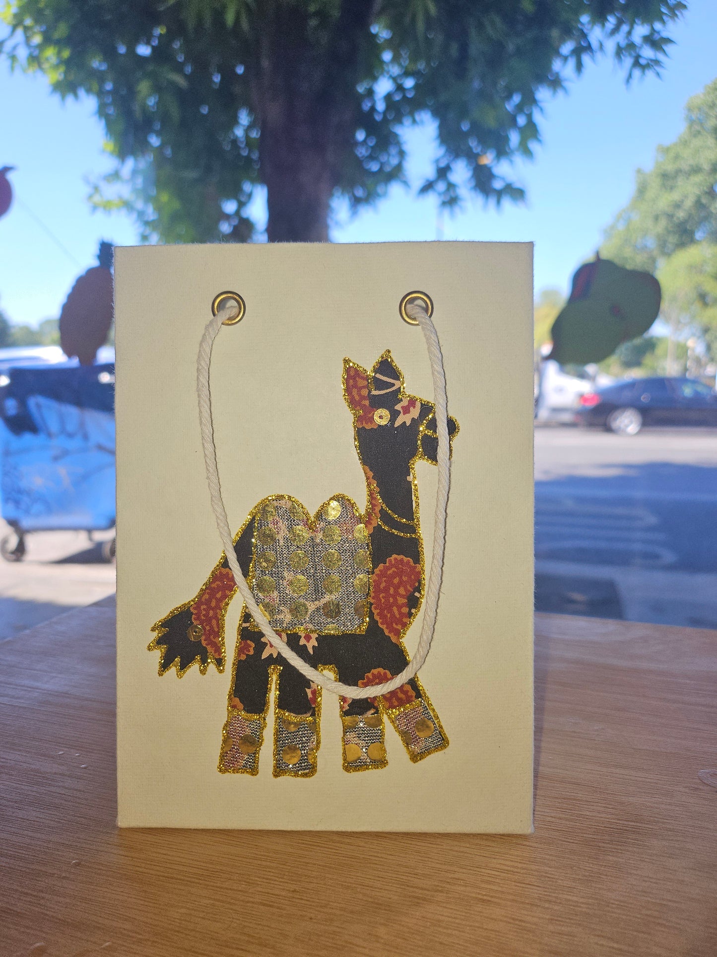 Decorative card with a camel made out of sari fabric design on a wooden surface with a blurred outdoor background.