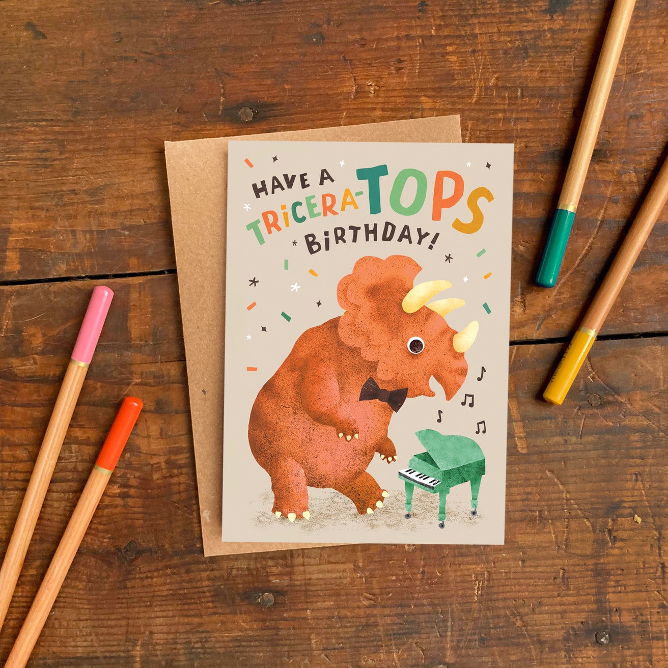 Birthday card with triceratops illustration on a wooden surface with pens.