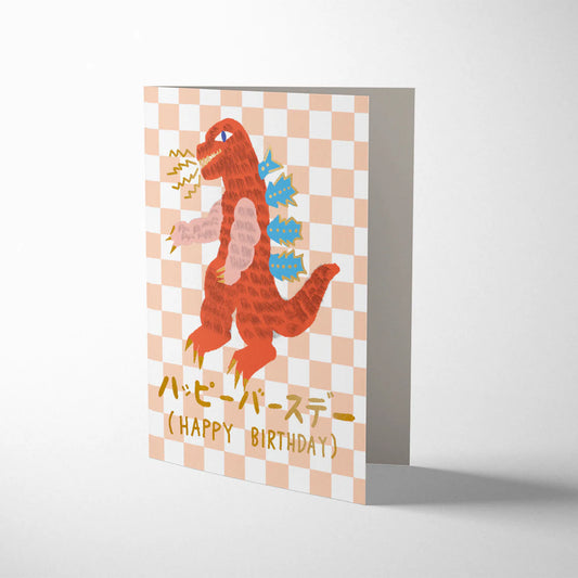 Birthday card with a monster illustration on a checkered. Kaiju Happy Birthday card by Carolyn Suzuki, featuring giant monster illustration, litho printed with gold foiling, kraft envelope, size 108mm x 140mm.background