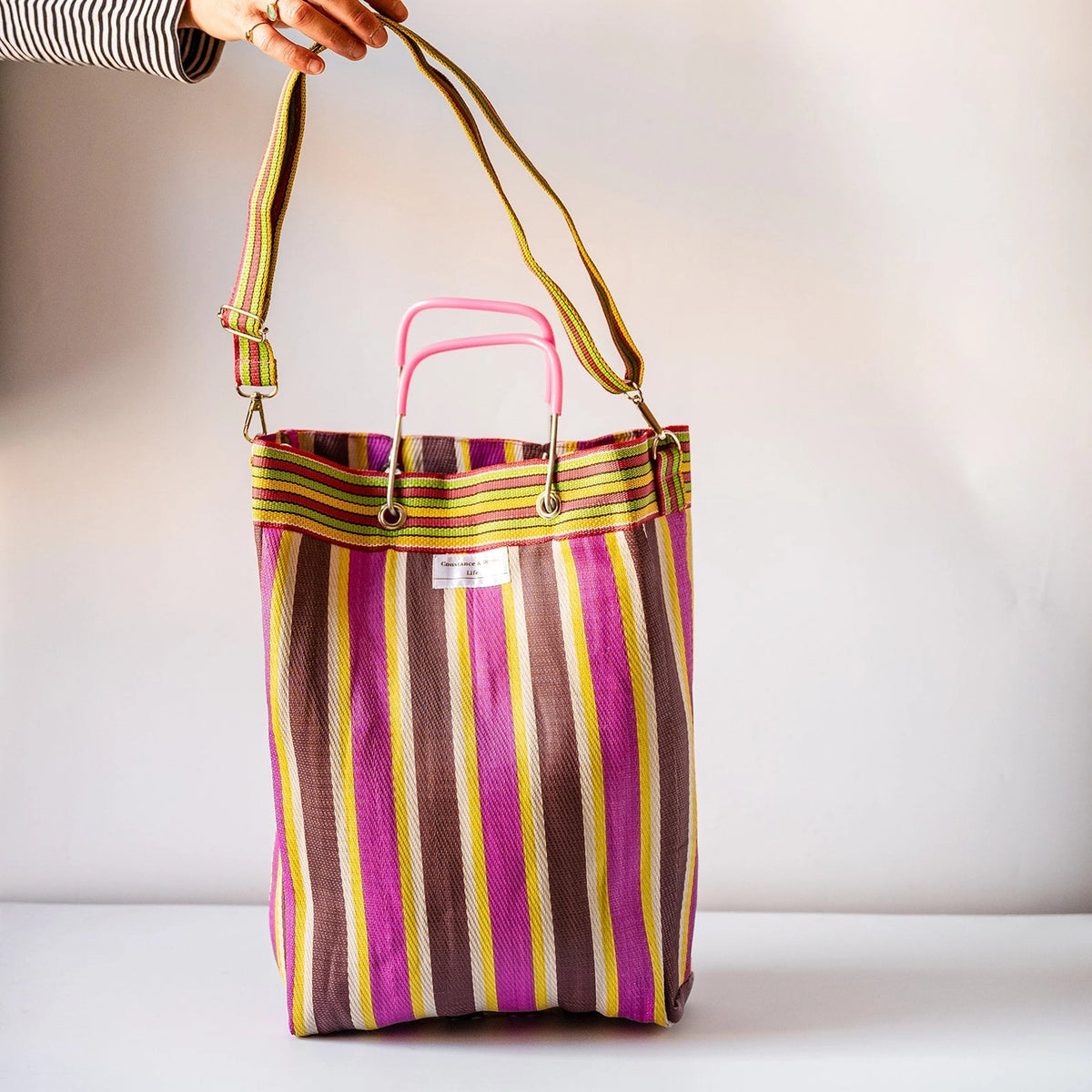 A striped tote bag with vertical pink, brown, yellow, and white stripes, featuring a pink top handle and a long adjustable shoulder strap held by a hand. The bag is set against a plain light gray background.