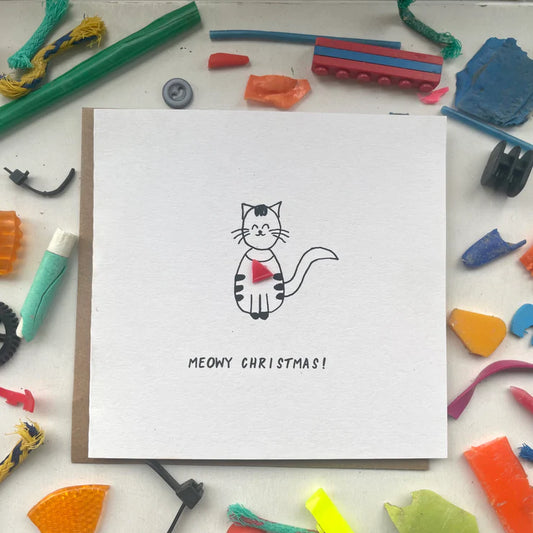 A handmade Christmas card with a drawing of a cat and the text 'Meowy Christmas!' on it, presented on a background of various colorful plastic pieces.