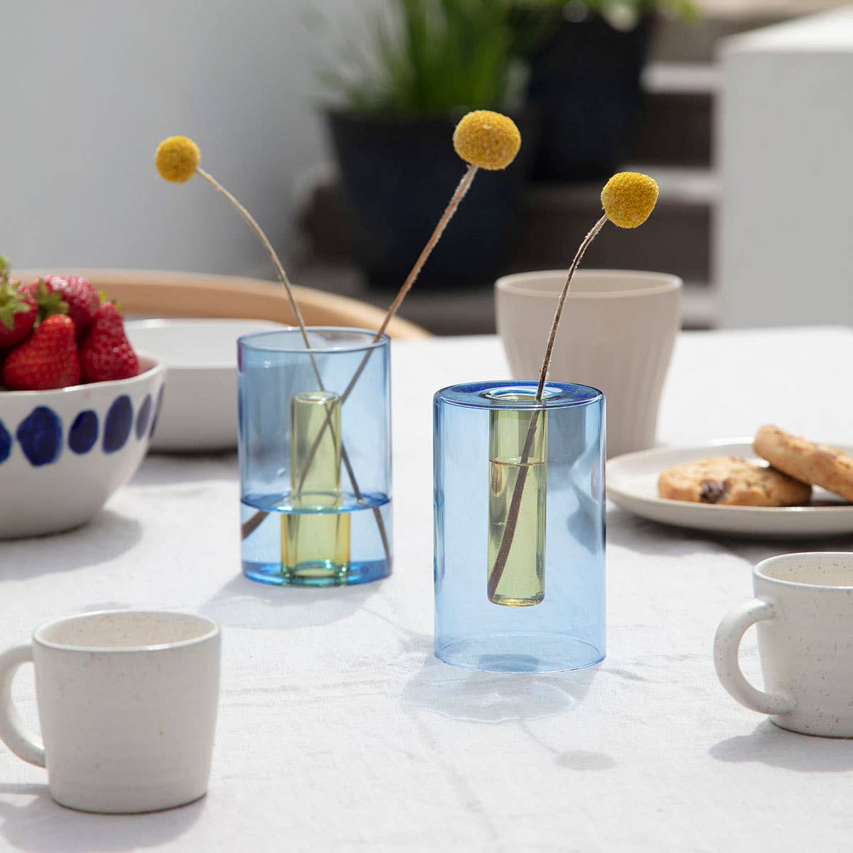 Two blue glass vases with floral arrangements on a table with a cup, bowl, and plate in the background.