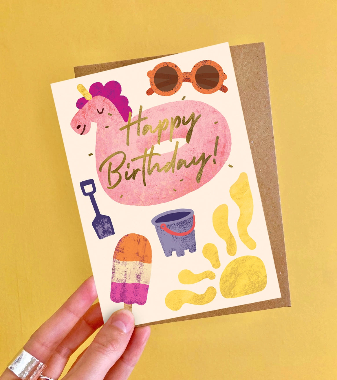 Kids birthday card with a pink unicorn inflatable, sunglasses, and beach-themed illustrations on a yellow background.