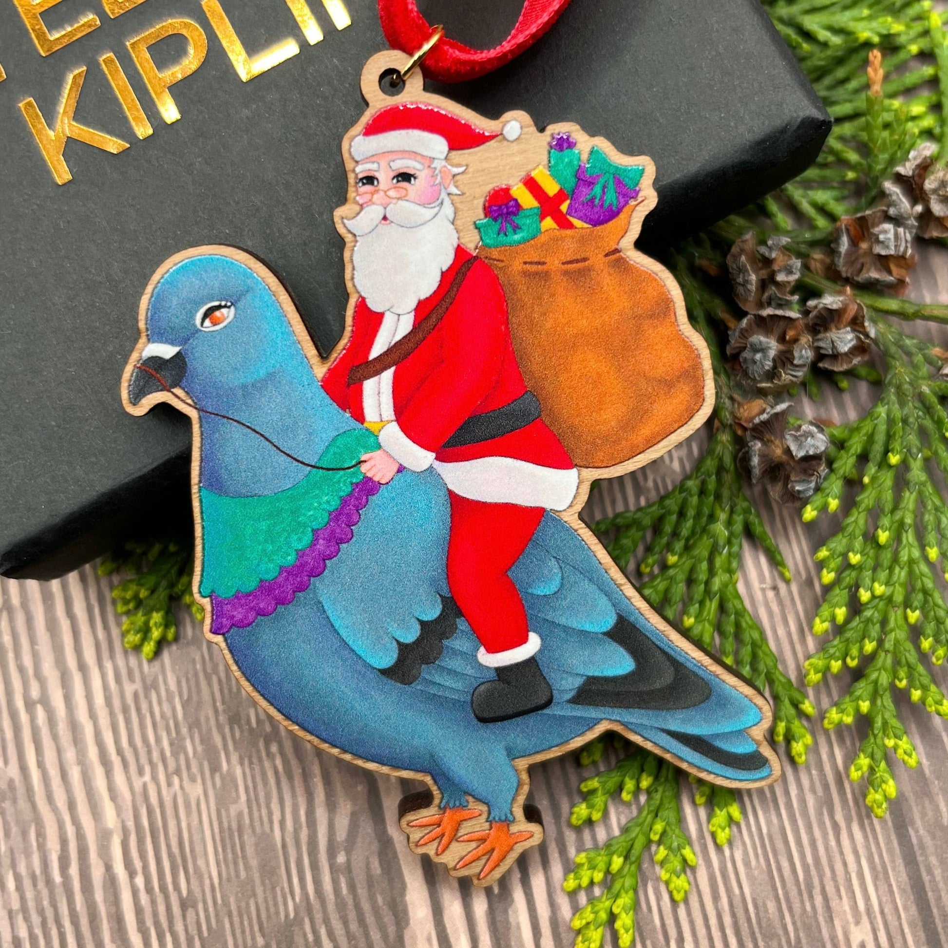 Santa Pigeon wooden Christmas tree decoration with Teddy & Kipling illustration, sustainably certified cherry wood, gold-plated ring, and red velvet ribbon.
