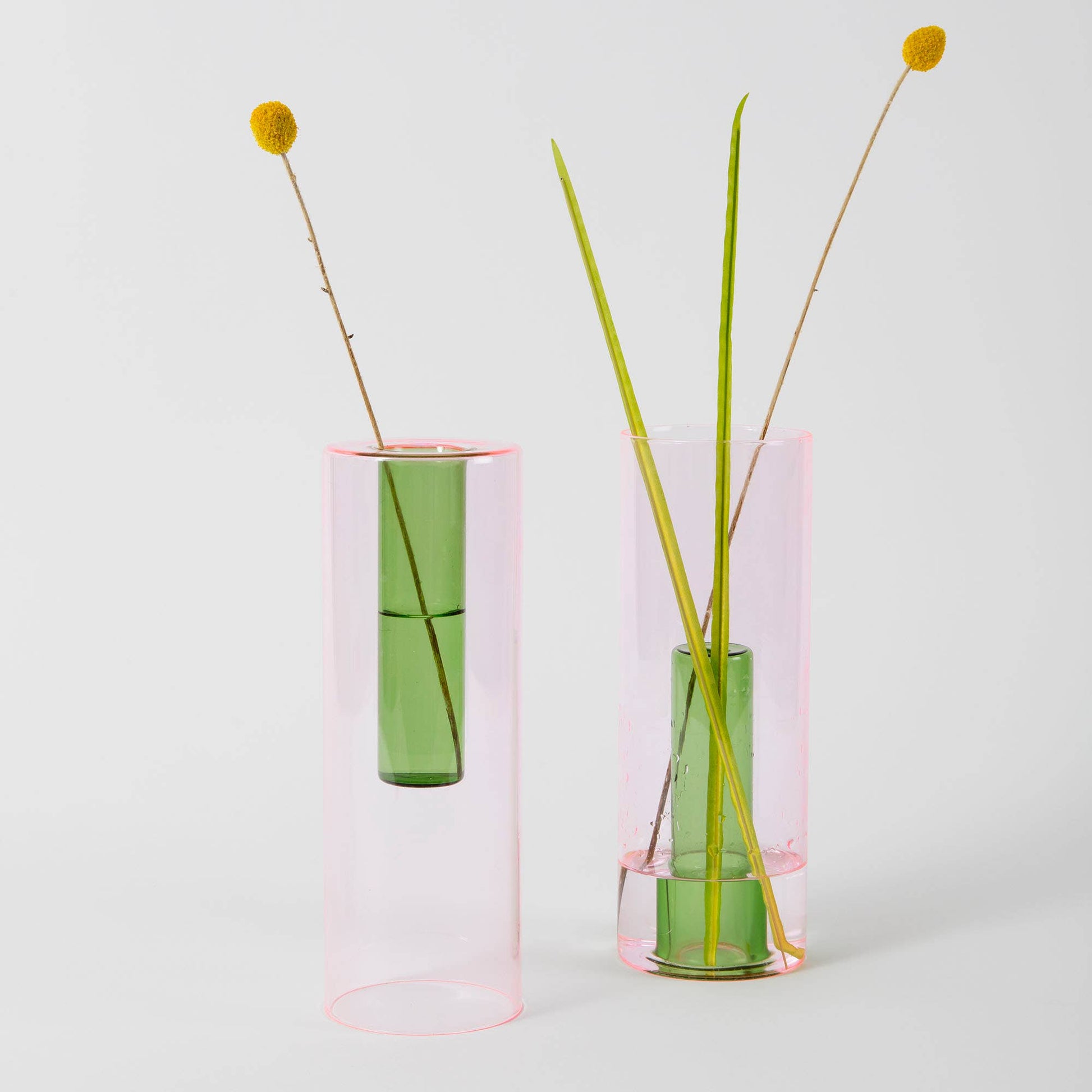 Two transparent pink vases with green stems and yellow flowers on a light gray background