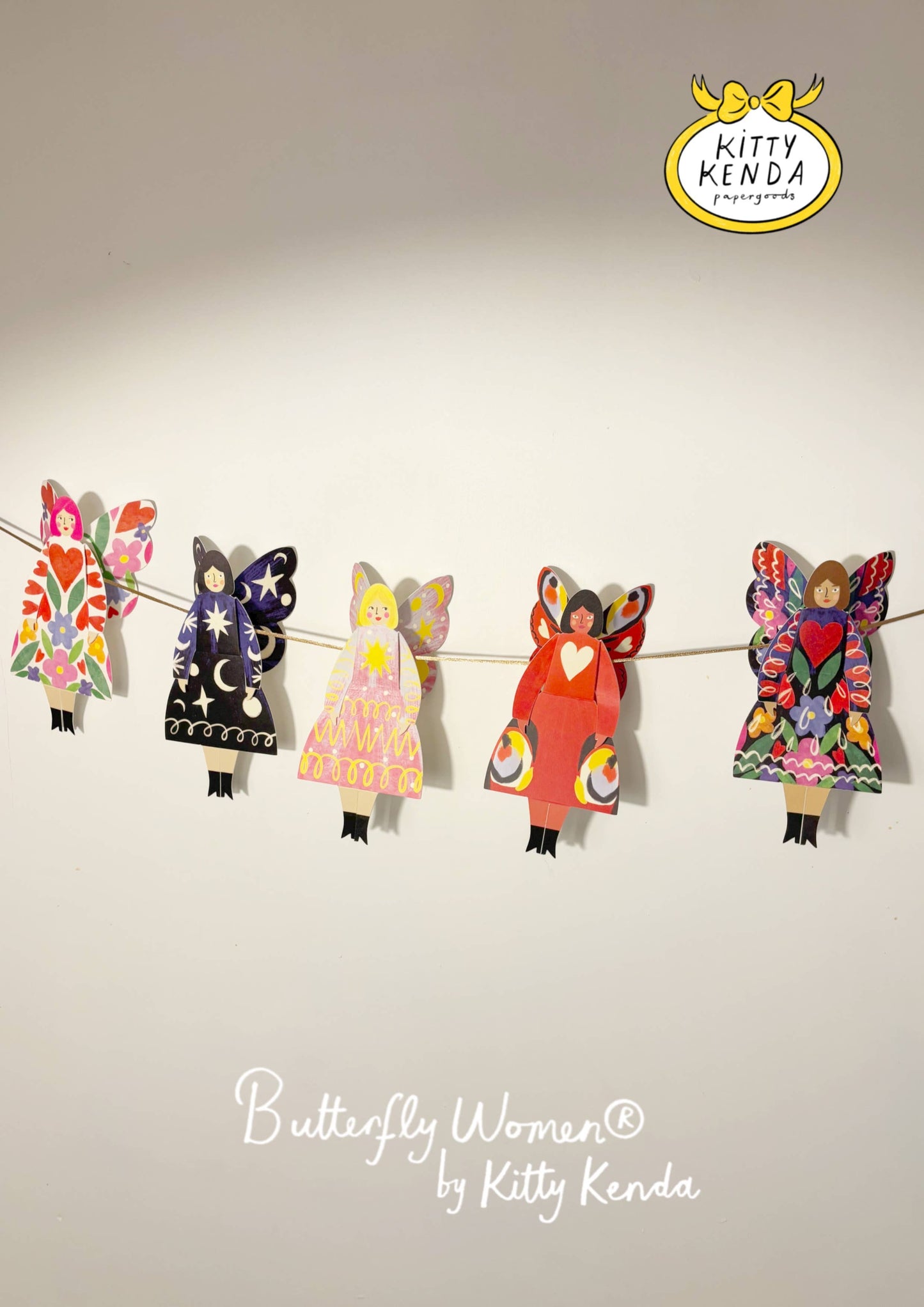 Colorful paper dolls of 'Butterfly Women' by Kitty Kenda hanging on a string against a plain background.