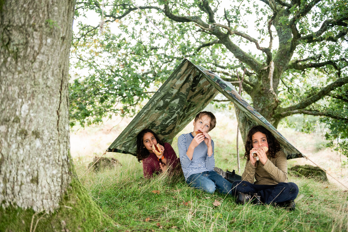 Children sitting under the camouflage tarpaulin which is weatherproof. Eating hot dogs.