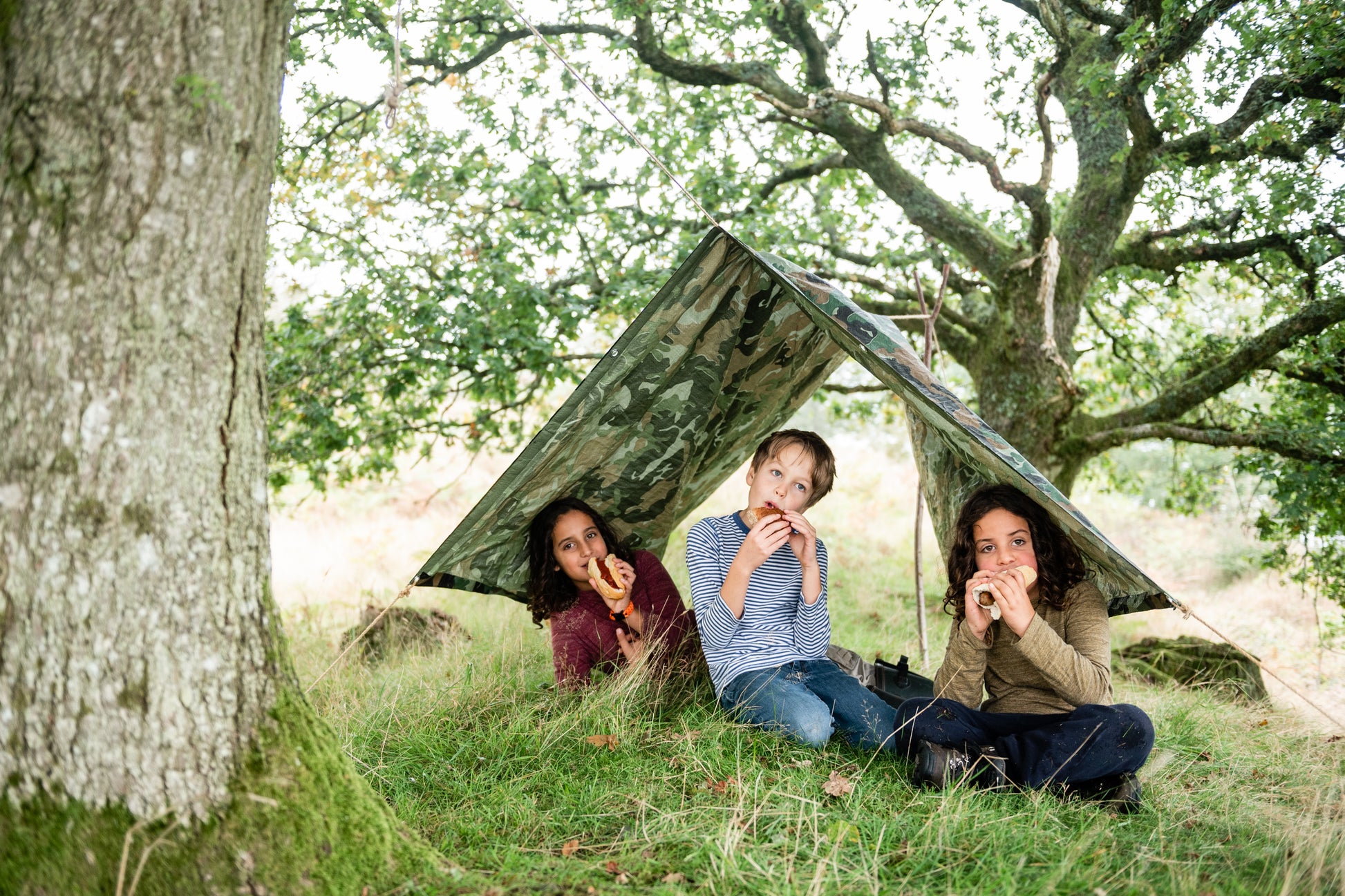 Children sitting under the camouflage tarpaulin which is weatherproof. Eating hot dogs.