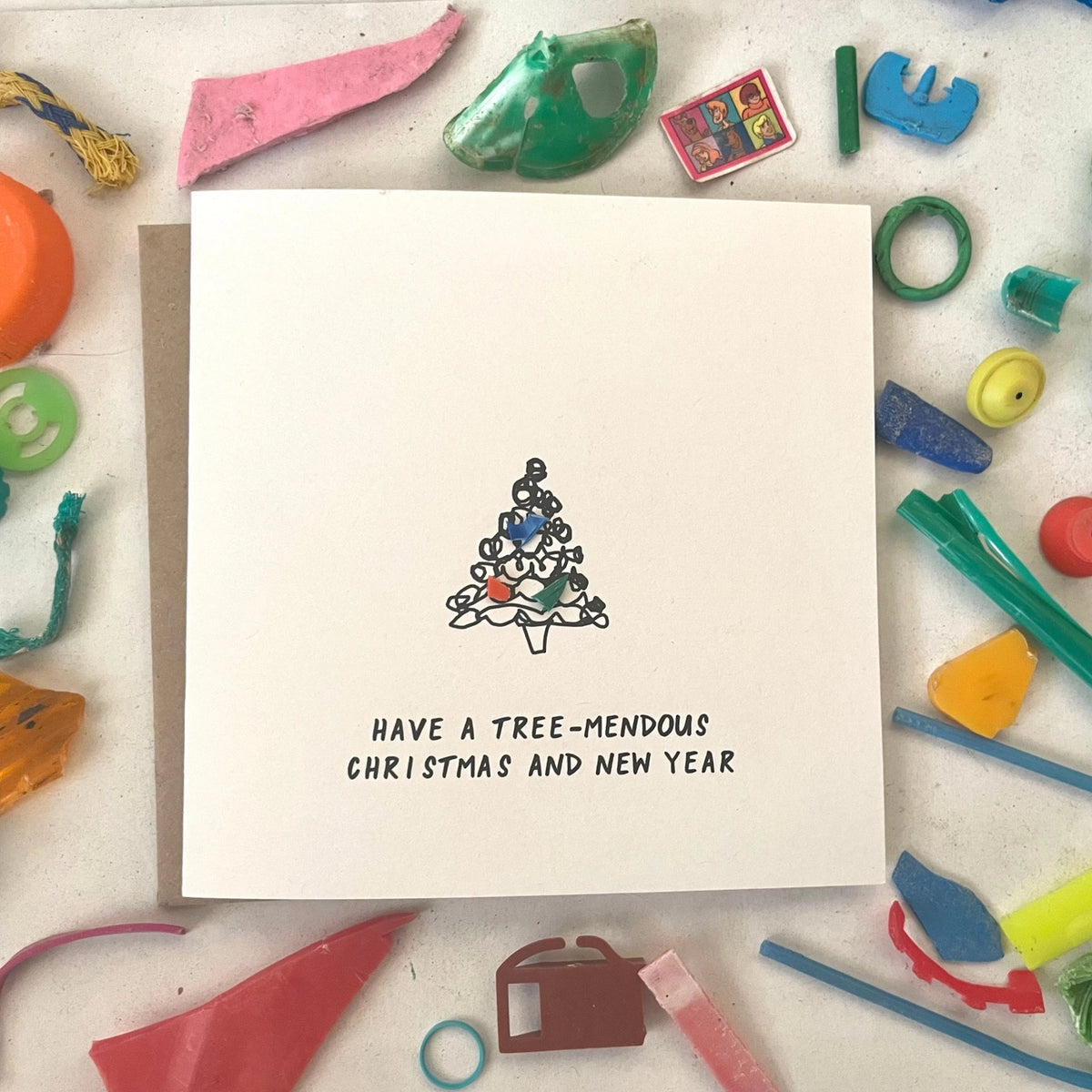 A square white greeting card with a small black-and-white Christmas tree illustration and the text 'HAVE A TREE-MENDOUS CHRISTMAS AND NEW YEAR' at the bottom, placed on a light surface surrounded by colorful craft scraps and small objects.