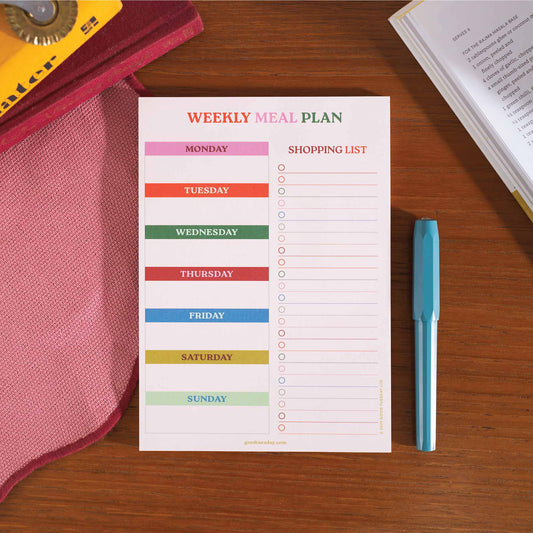 A5 size meal planner and shopping list pad with colorful headers for each day of the week, placed on a wooden surface with a pen beside it.
