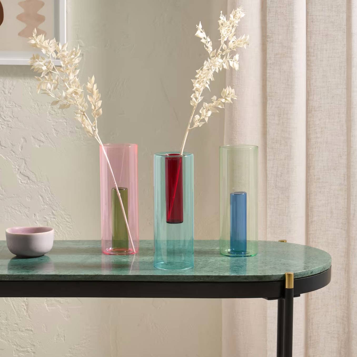 Three colorful vases with dried plants on a green table against a neutral wall.