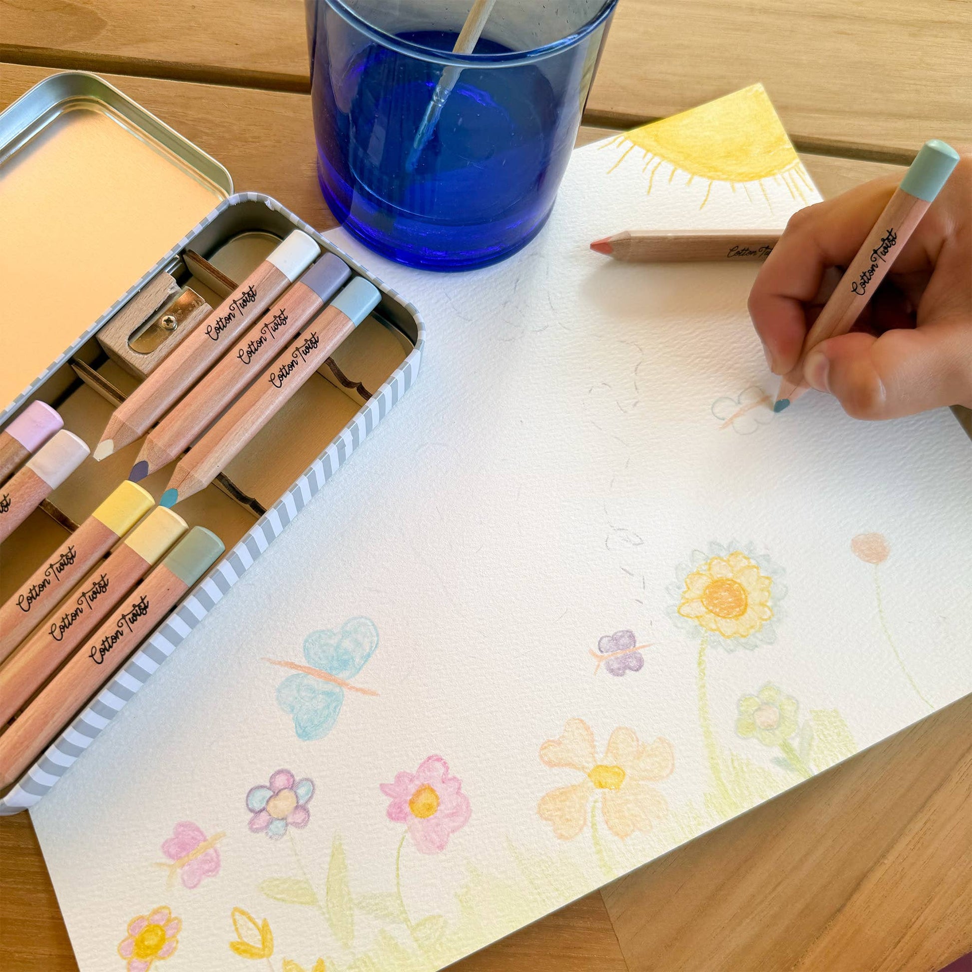 Colouring with pastel-coloured pencils on a piece of paper with floral designs, next to a blue glass and pencil sharpener.