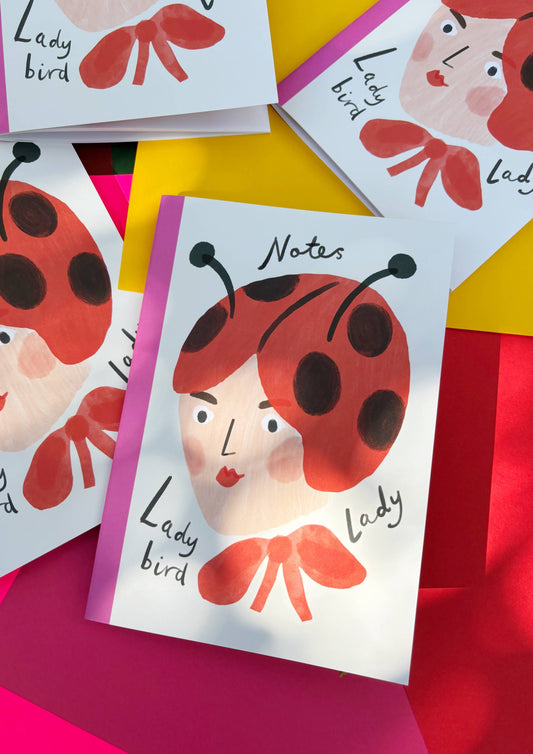 Illustration of a lady's head adorned with a ladybird hat and big red bow. With the words Notes at the top and  Lady bird, Lady across the bottom. The Notebook is bound with a pink edge and has plain paper for notes and sketching.