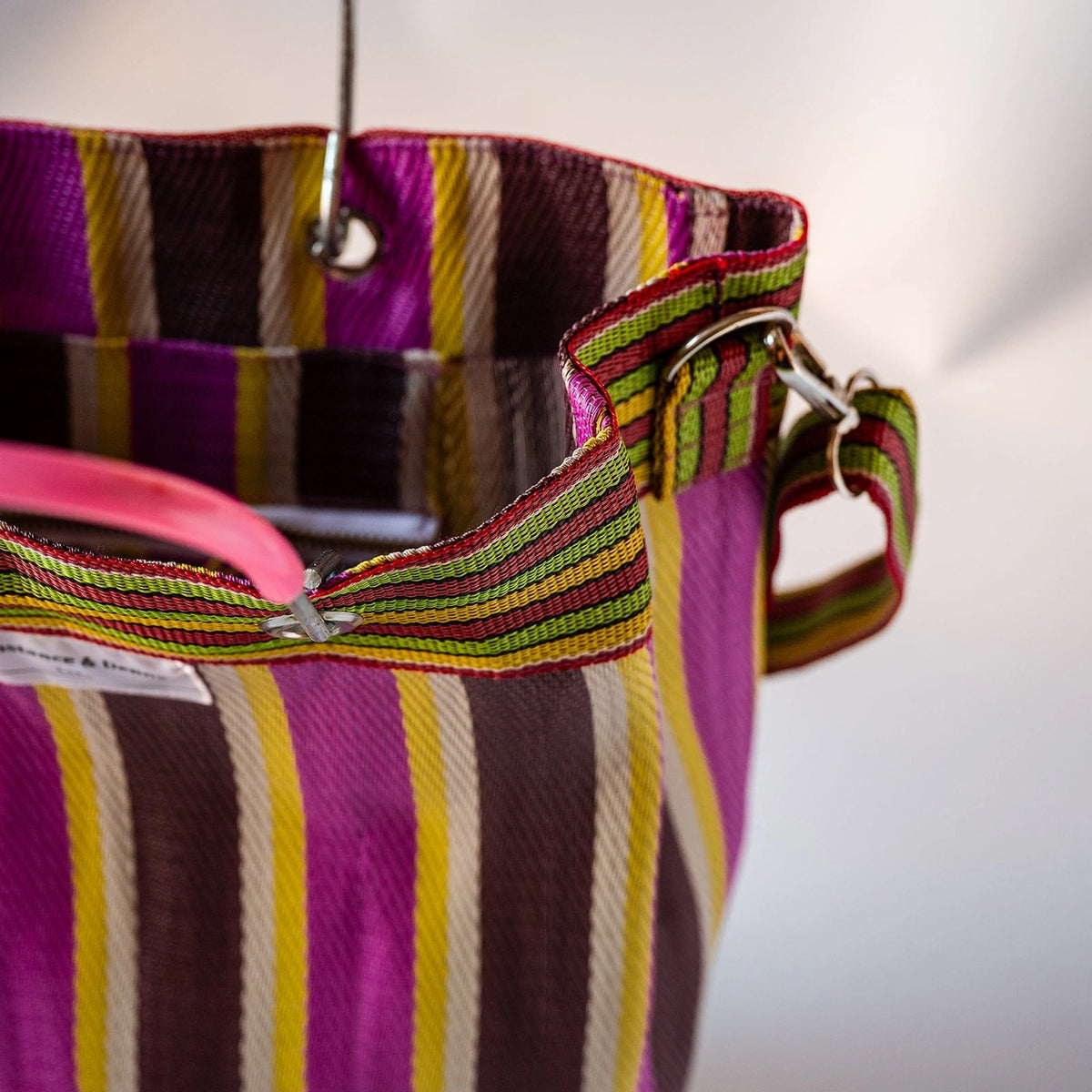 Close-up of a woven tote bag with vertical purple, brown, yellow, and white stripes, featuring a pink handle and a matching striped shoulder strap with metal hardware.