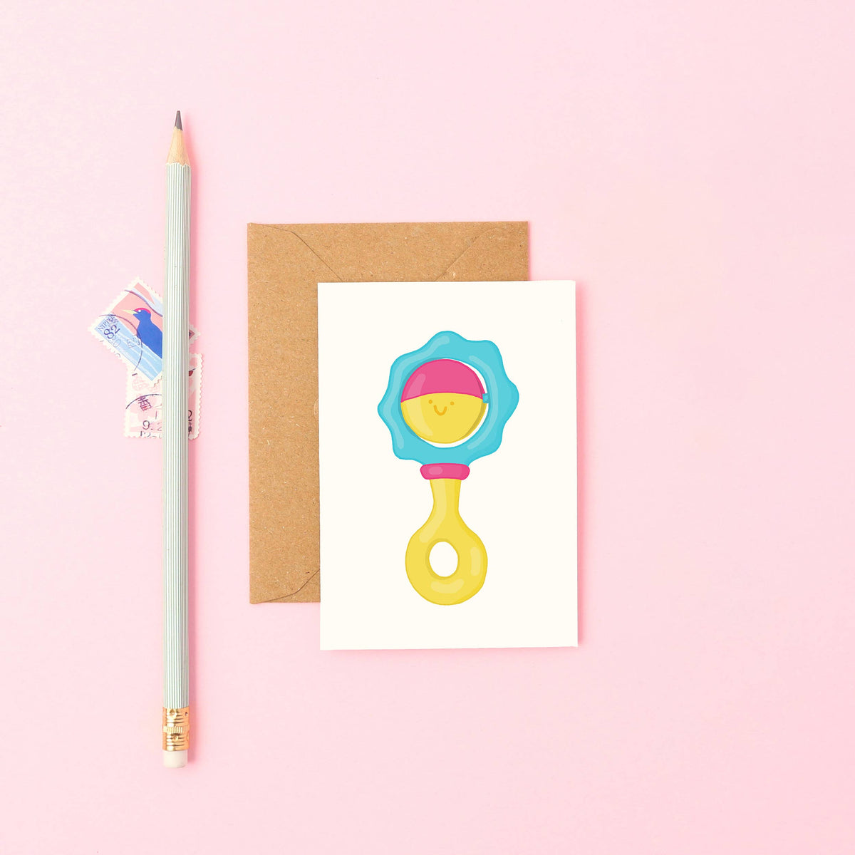 Hand-drawn baby rattle mini card with a cute face in blue, pink, and yellow