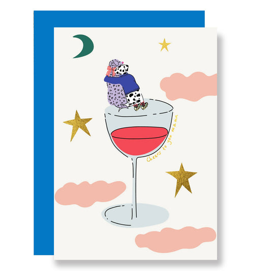 Cheers to you mum in gold foil. Two dogs sit cuddling with their backs to at the top of a glass of red wine, which is surrounded by the moon, stars and pink fluffy clouds.