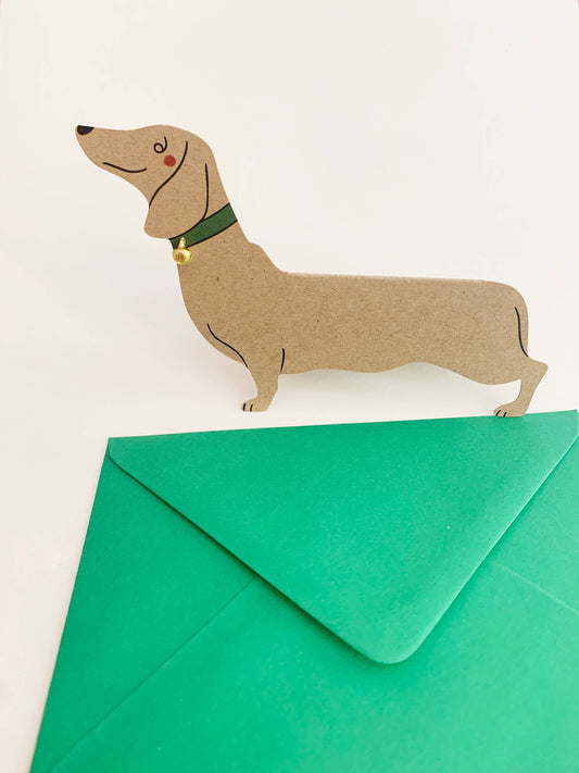 This adorable standing dachshund card is perfect for any occasion! Birthdays, thank you's or I love you's. Hand finished with a tiny gold bell, this recycled 300gsm card is exclusive to Kitty Kenda, designed in-house and manufactured in the UK from sustainable materials with a bright green envelope.