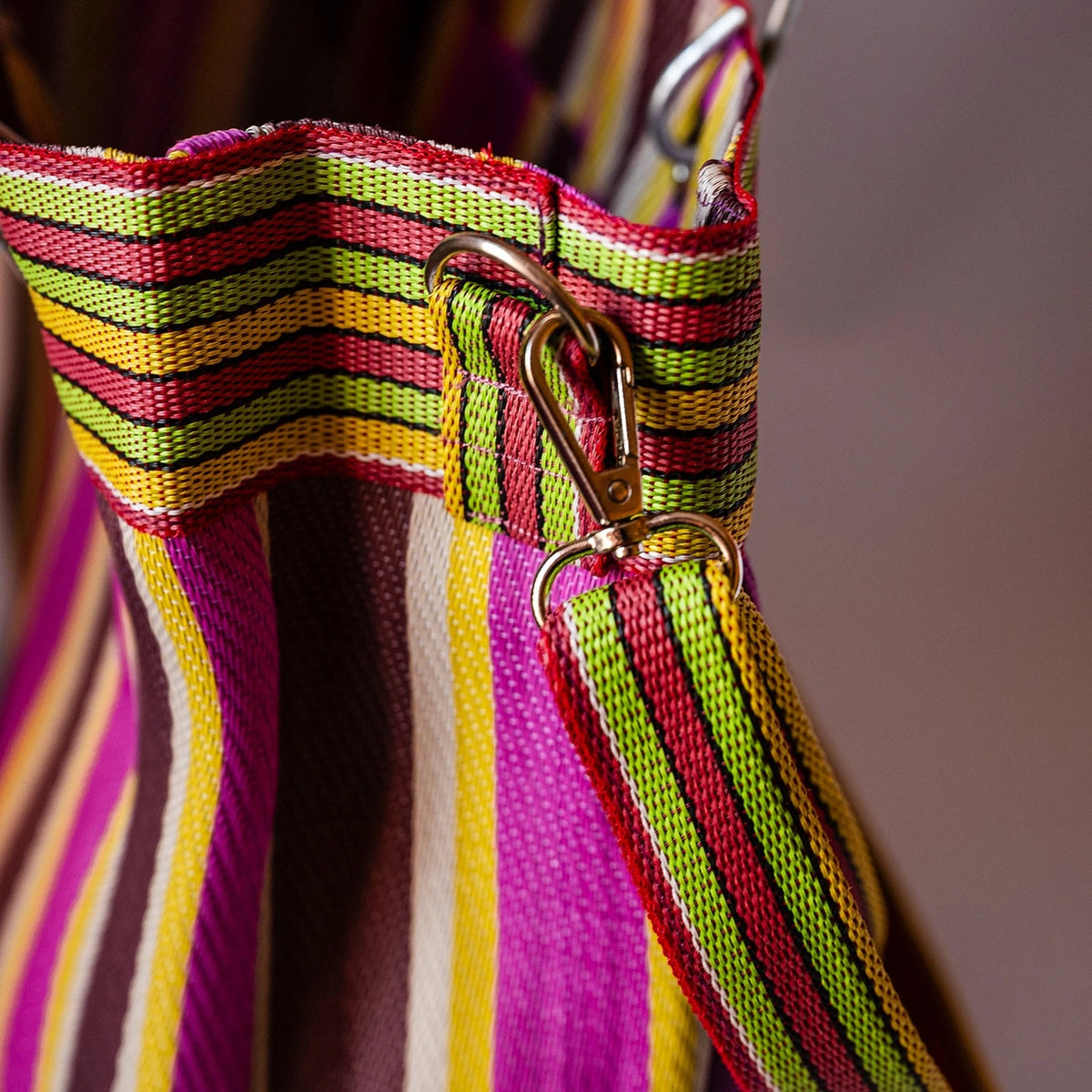 Close-up of a striped fabric bag or tote with a metal clasp, showing bright pink, green, yellow, red, and white woven stripes against a soft blurred background.