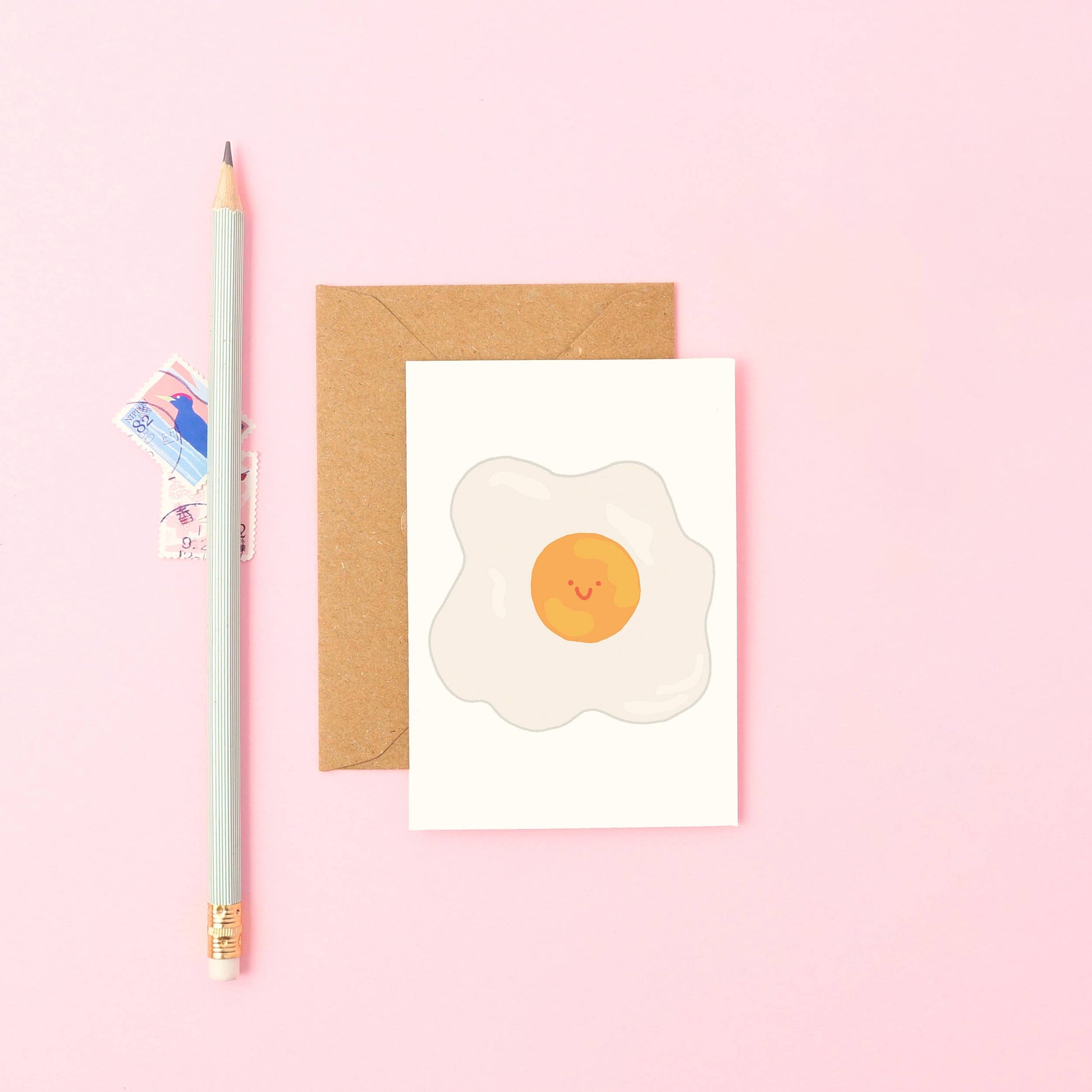 Card with a smiley face egg design on a pink background with a pencil and envelope.