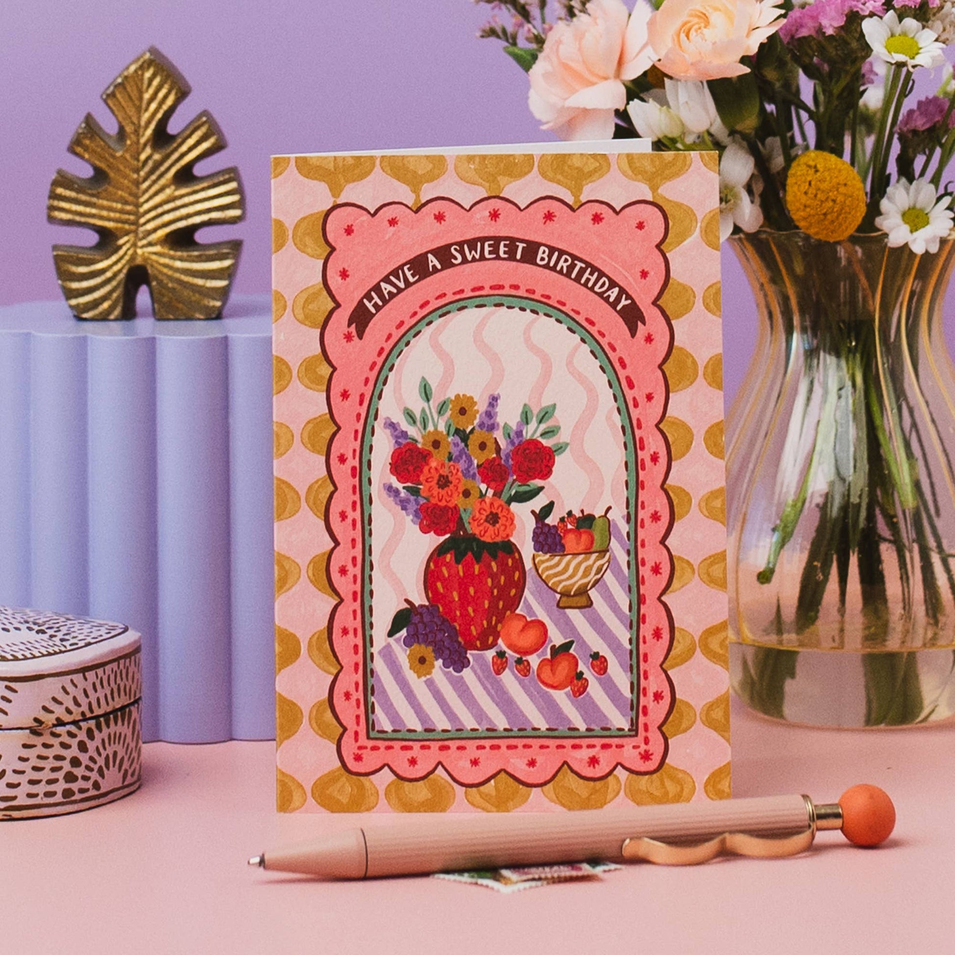 Hand drawn lettering reads 'Have a sweet birthday'. Part of the on trend Harlequin range, which features contemporary vases of flowers and fruit in a patterned tablescape. A6 size illustrated greeting card with tile pattern and vase of flowers and fruit illustration, printed onto premium textured card with a kraft envelope.