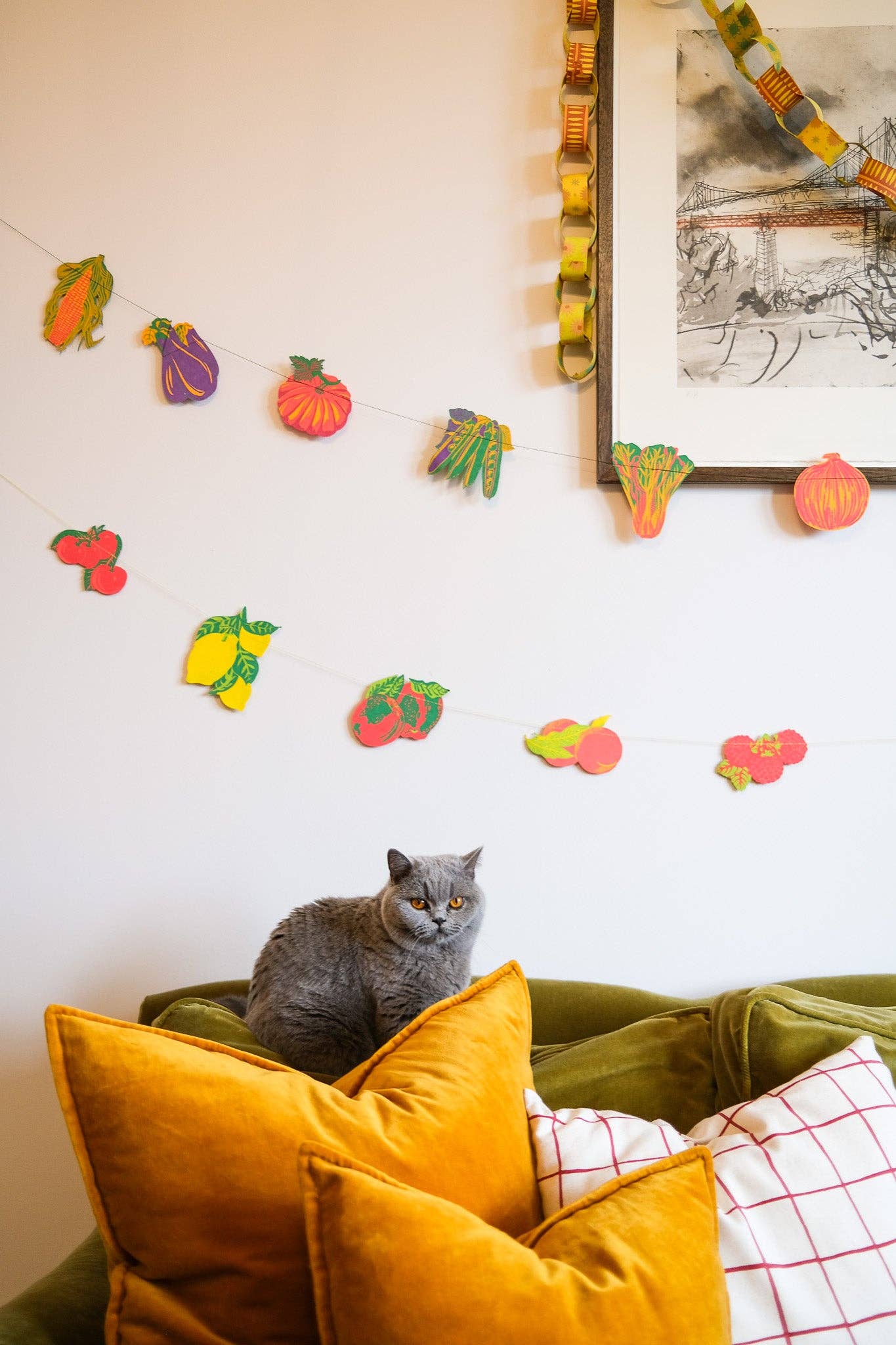 Colourful Fruit Sewn Garland hung above a sofa with a cat on cushions, showcasing vibrant fruit designs as playful home decor.