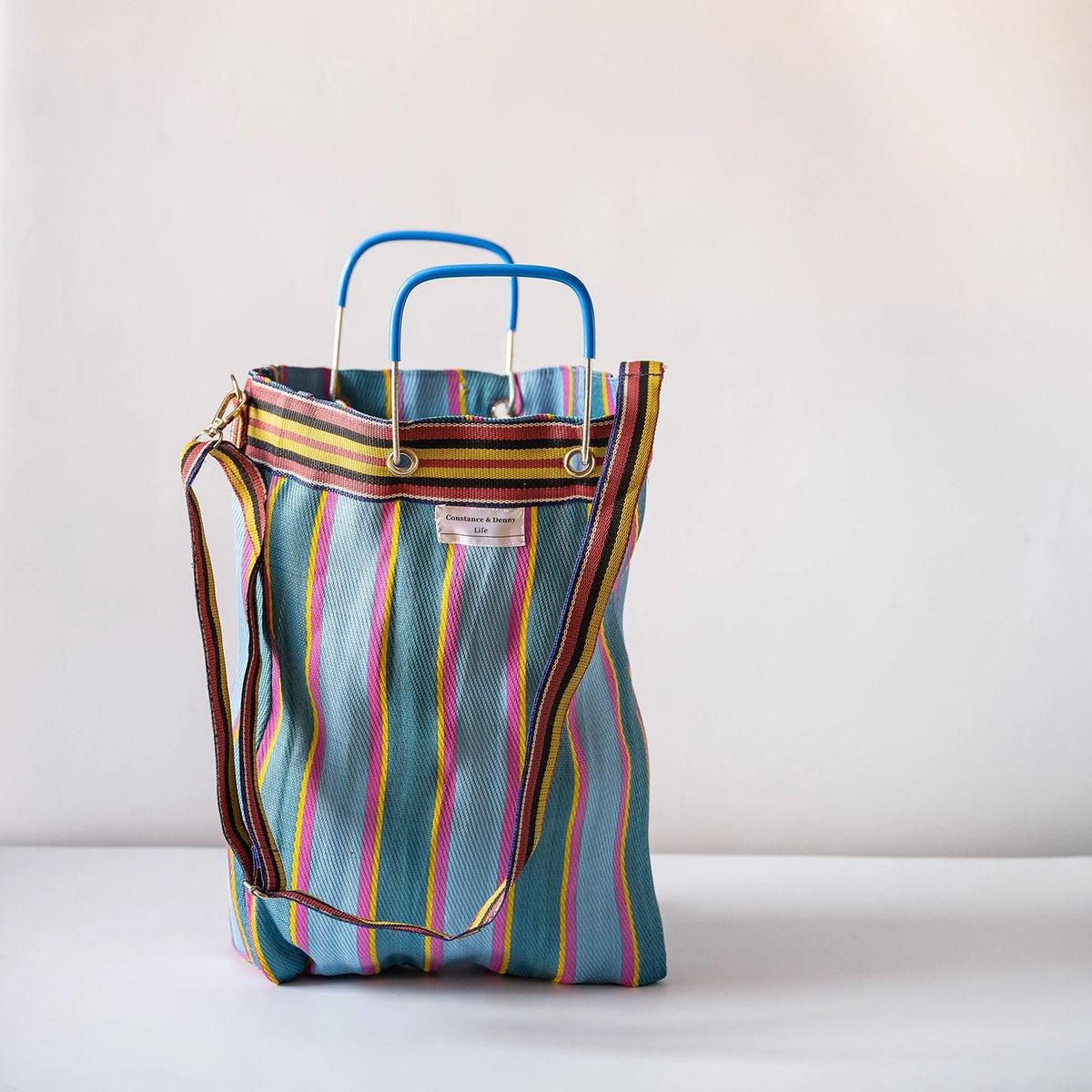 A striped tote bag with a teal-blue base, vertical stripes in pink, yellow, orange, and green, featuring blue double top handles and a brown shoulder strap, set against a plain light background.