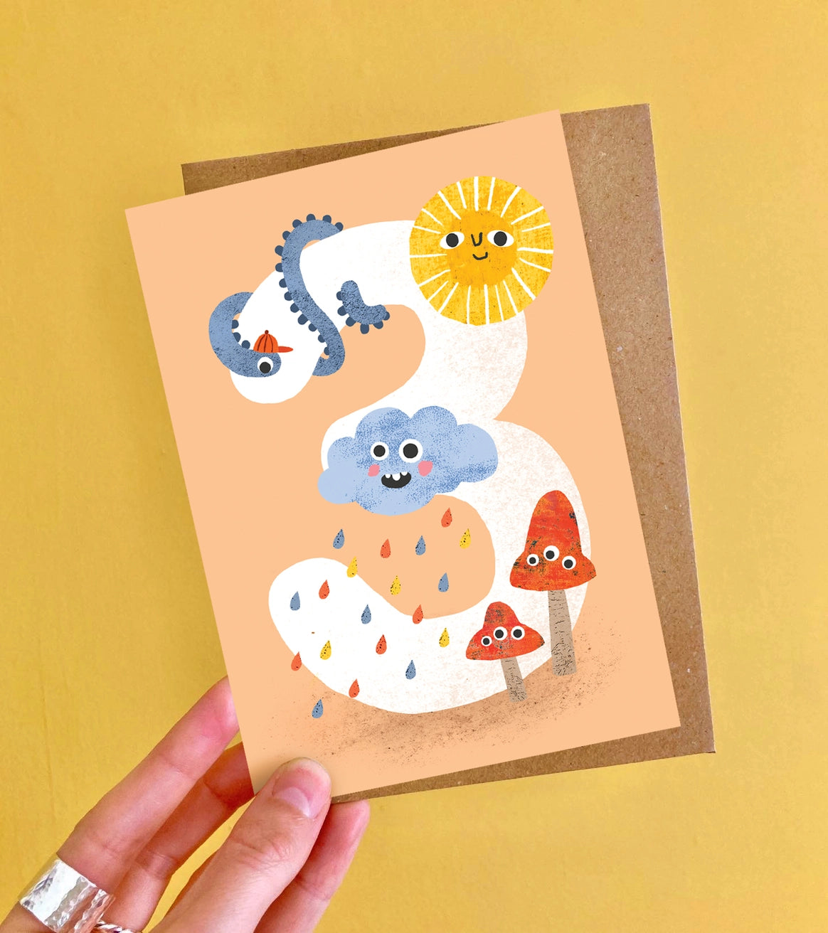 Age 3 birthday card with colourful cloud, sun, and mushroom illustrations on a peach background. Made in the UK.