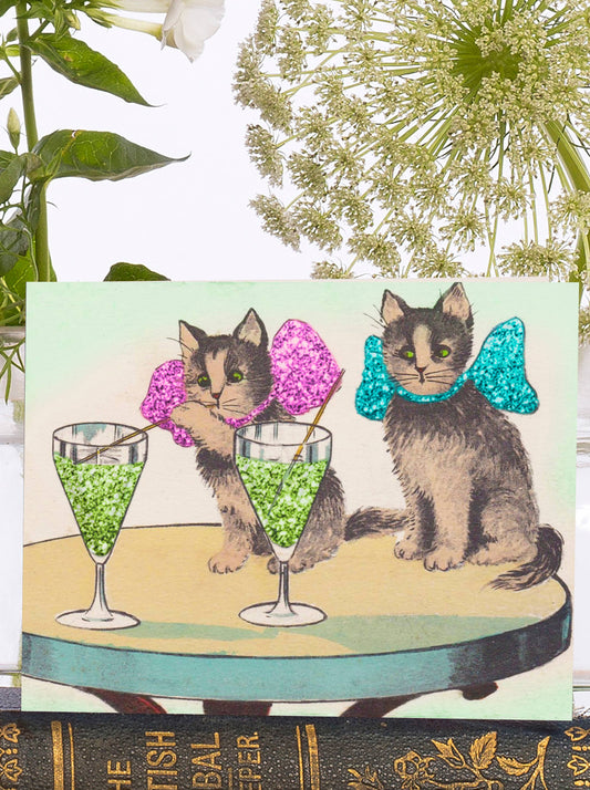Festive kittens, adorned with glittery pink and blue bows, sip on a Christmas drink in this vintage-inspired illustration.